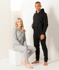 Two individuals in matching solid-colored, zip-up hooded onesie pajamas (SF470 by SF) stand against a plain backdrop; the one in black smiles while standing, and the one in gray sits barefoot on a white block.