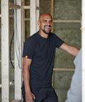 A person in a black Scruffs SH001 t-shirt stands inside a building under construction, smiling and interacting with someone off-frame. Wooden framing and insulation are visible in the background.