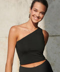 A person stands outside by a light-coloured wall, smiling at the camera with hair tied back, wearing the SF Women's one-shoulder top in black and matching trousers.