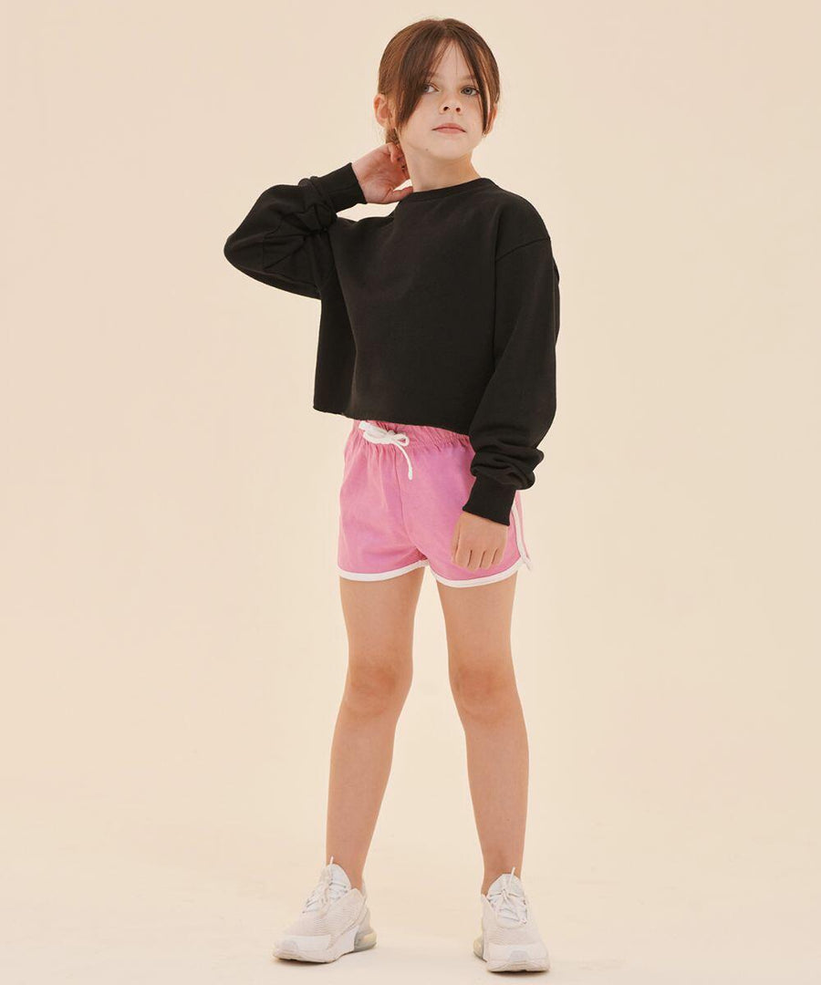 A young person stands against a plain background wearing a black long-sleeve top, pink shorts, and SF SM069 white sneakers, with one hand touching their neck.