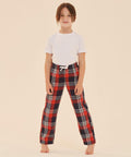 A child with long hair stands barefoot, wearing a white t-shirt and red and black plaid SF Minni SM083 pajama pants against a plain beige background.