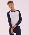 A young person wearing an SF SM271 long-sleeve raglan shirt with navy sleeves and a white body, and dark pants, stands against a plain pink background.