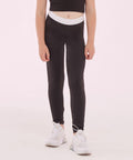 A person wearing a black SM426 crop top, black SF Minni leggings with a white waistband, and white sneakers stands against a light pink background.
