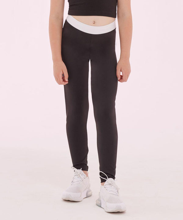 A person wearing a black SM426 crop top, black SF Minni leggings with a white waistband, and white sneakers stands against a light pink background.