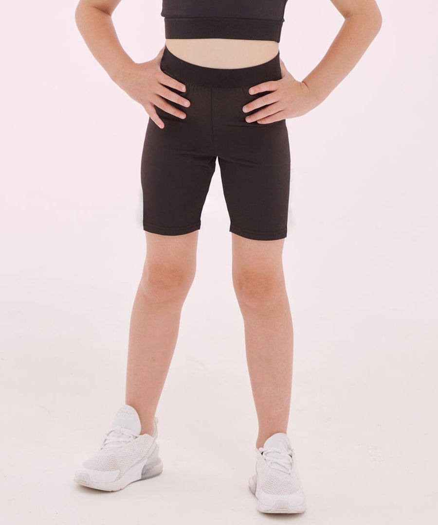 A person standing with hands on hips, wearing a black top and black SF Minni SM427 shorts, white socks, and white sneakers, against a plain light background. The person's torso is not visible.