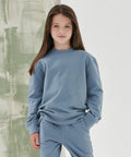 A young girl with long brown hair stands wearing a light blue SM530 sweatshirt and matching pants from SF Minni. She has one hand in her pocket and is standing against a green and white background.