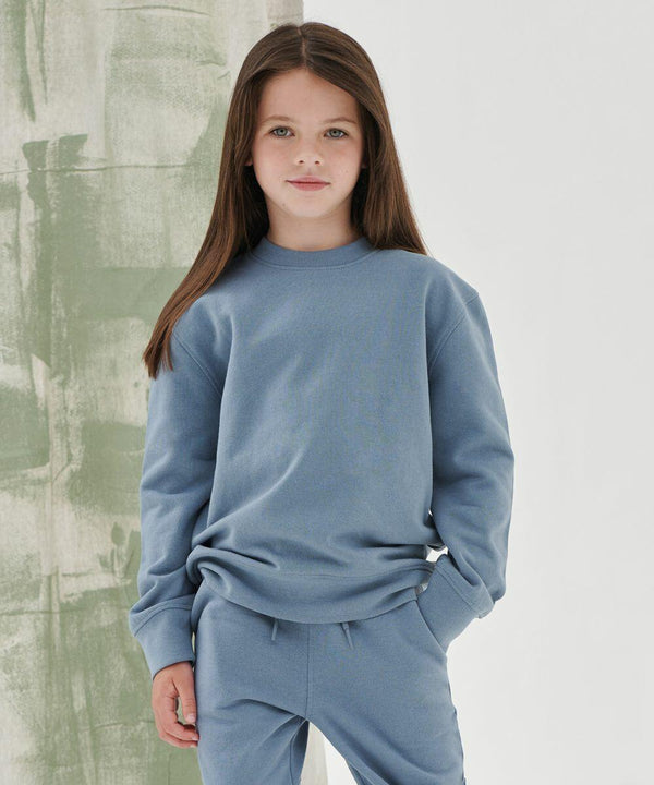 A young girl with long brown hair stands wearing a light blue SM530 sweatshirt and matching pants from SF Minni. She has one hand in her pocket and is standing against a green and white background.