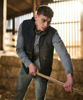 Person in a barn cleaning with a Regatta SN104, wearing a black vest over a blue shirt and jeans. Hay bales and sunlight through wooden slats are visible in the background.