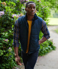A person walks on a path through a garden, wearing a blue quilted vest (SN133 by Regatta) over a yellow shirt and blue checkered shirt.
