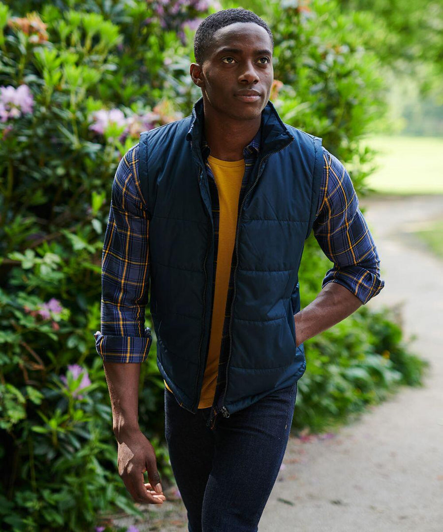 A person walks on a path through a garden, wearing a blue quilted vest (SN133 by Regatta) over a yellow shirt and blue checkered shirt.