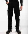 A person is wearing black cargo pants with multiple pockets, a black long-sleeve top, and Kariban SP105 shoes with orange accents. The background is plain white.