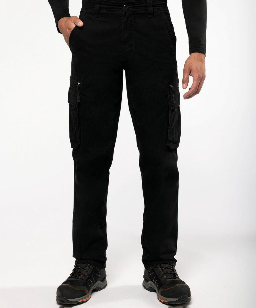 A person is wearing black cargo pants with multiple pockets, a black long-sleeve top, and Kariban SP105 shoes with orange accents. The background is plain white.