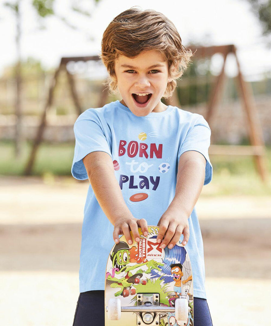 A young boy with light brown hair and a blue "Born to Play" Fruit of the Loom SS031 T-shirt smiles while holding a colorful skateboard. A playground structure is visible in the blurred background.