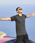 A person wearing sunglasses and a dark Fruit of the Loom SS034 t-shirt stands with arms outstretched, smiling, in front of a scenic view of the ocean and cliffs.