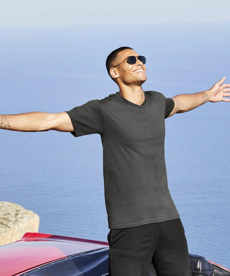 A person wearing sunglasses and a dark Fruit of the Loom SS034 t-shirt stands with arms outstretched, smiling, in front of a scenic view of the ocean and cliffs.