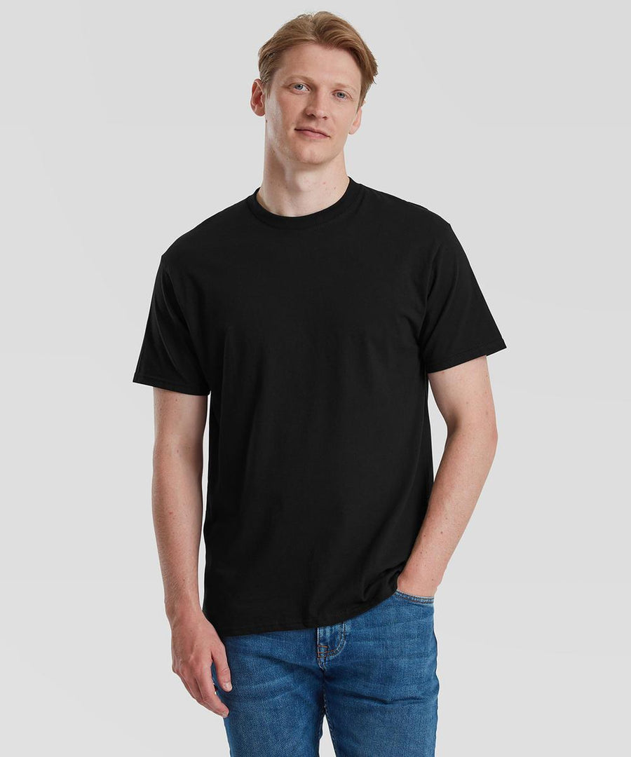 A person with fair skin and blond hair wears a Fruit of the Loom Pure cotton T-shirt in black and blue jeans, standing against a plain light grey background with one hand in their pocket.