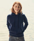 Person with long red hair smiling and wearing a navy blue Fruit of the Loom SS038 hoodie stands outdoors near a body of water.