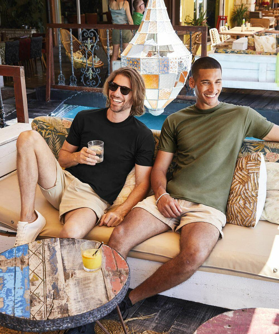Two men sitting on a couch outdoors, one in a black Fruit of the Loom SS044 shirt holding a drink and the other in a green shirt. They are both smiling and appear to be relaxed. A colorful lamp hangs in the background.