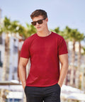A person wearing Fruit of the Loom SS048 sunglasses, a red t-shirt, and dark shorts stands outdoors with palm trees and a building blurred in the background.