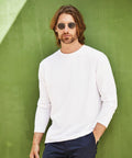 A man with shoulder-length hair and sunglasses is wearing a white long-sleeve SS069 shirt from Fruit of the Loom and dark pants, standing against a green wall.