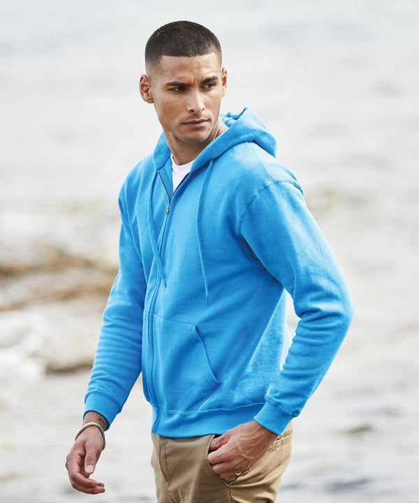 Person in a blue hoodie and tan pants stands near a body of water, looking to the side while wearing a Fruit of the Loom SS222.