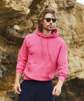 A person with long hair and sunglasses wears a pink Fruit of the Loom SS224 hoodie and dark pants, standing outdoors near a rocky terrain.