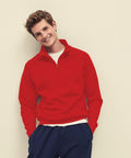 A person wearing a red Fruit of the Loom SS230 sweater and blue pants stands against a light-colored wall, smiling with hands in pockets.