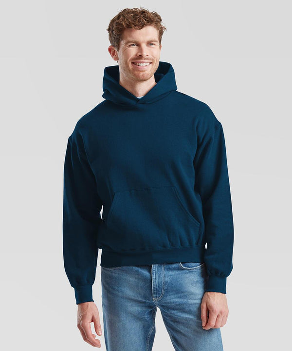 A man stands facing forwards against a plain light grey background, smiling with one hand relaxed at his side, wearing a dark blue Fruit of the Loom Supercotton hooded sweatshirt and light blue jeans.