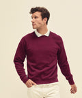 A man in a burgundy Fruit of the Loom SS270 sweatshirt and white collared shirt looks to the side. He has short, curly brown hair and stands against a plain, light-colored background.