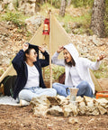Two people sit laughing in front of a small tent in a wooded area. They are wearing jeans and Fruit of the Loom SS312 hoodies, with a lantern, a pile of rocks, and a camping stove in the foreground.