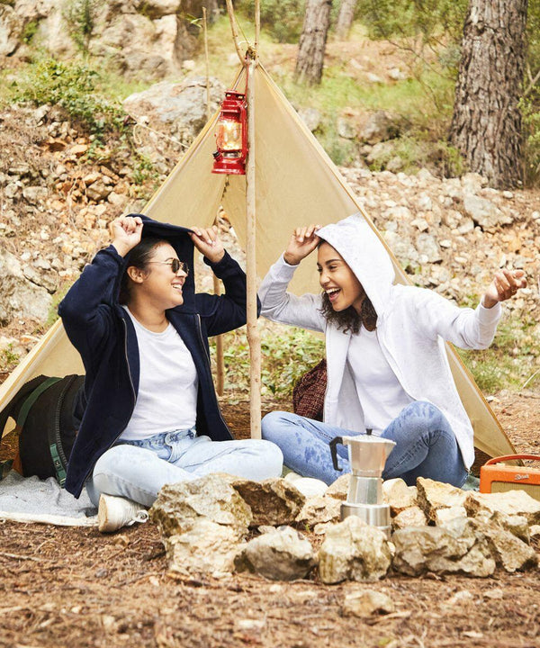 Two people sit laughing in front of a small tent in a wooded area. They are wearing jeans and Fruit of the Loom SS312 hoodies, with a lantern, a pile of rocks, and a camping stove in the foreground.