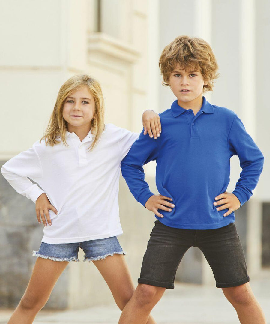Two children stand outdoors, smiling confidently. The girl on the left wears a white long-sleeve shirt and denim shorts. The boy on the right wears an SS320 blue long-sleeve shirt by Fruit of the Loom and black shorts.