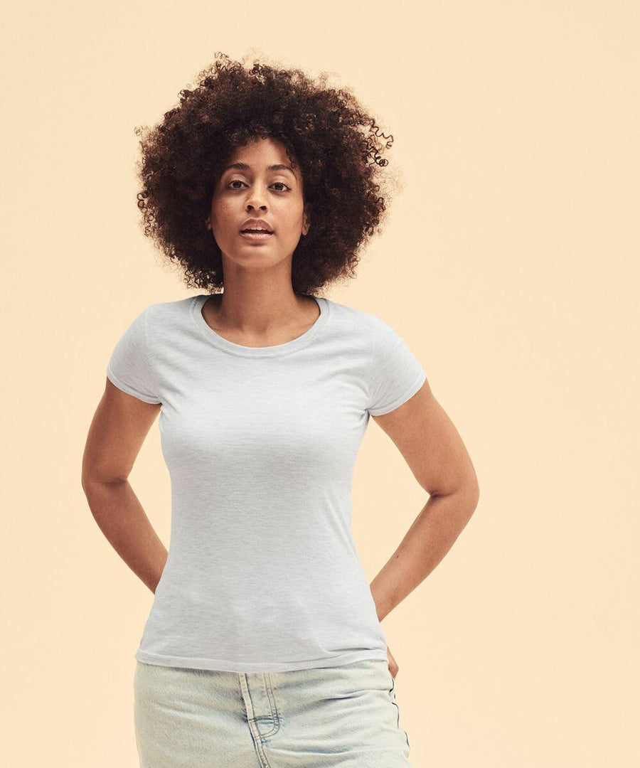 A person with curly hair wearing a light blue fitted Fruit of the Loom SS424 and jeans stands against a beige background.