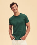 A man smiles while standing with his hands in his pockets. He is wearing a dark green Fruit of the Loom SS430 t-shirt and off-white pants against a plain beige background.