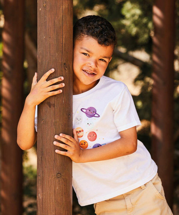 A young child wearing a white Fruit of the Loom SS436 t-shirt with colorful planet illustrations leans against a wooden post outdoors, smiling.