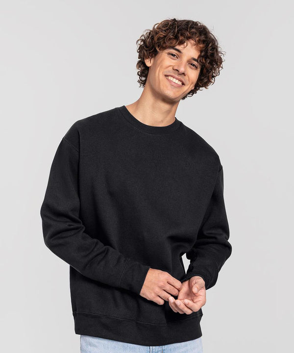 A person with curly hair stands against a plain background, smiling with hands clasped, wearing the Fruit of the Loom Iconic premium set-in sweatshirt in black and light blue jeans.