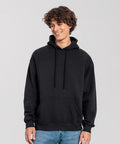 A person with curly hair is standing and smiling, wearing a Fruit of the Loom Iconic premium hooded sweatshirt in plain black with a front pocket, paired with blue jeans, against a light grey background.