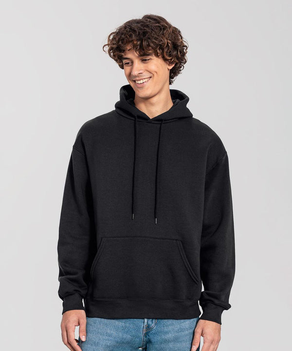 A person with curly hair is standing and smiling, wearing a Fruit of the Loom Iconic premium hooded sweatshirt in plain black with a front pocket, paired with blue jeans, against a light grey background.