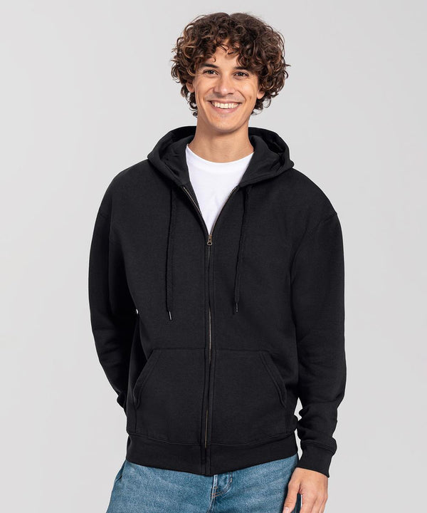 A person with curly hair smiles while wearing the Fruit of the Loom Iconic premium hooded sweat jacket in black, paired with a white T-shirt and blue jeans, standing against a plain light grey background.