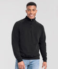 A person stands facing the camera, smiling, wearing a Fruit of the Loom Iconic premium zip-neck sweatshirt in black and light blue jeans against a plain light grey background.