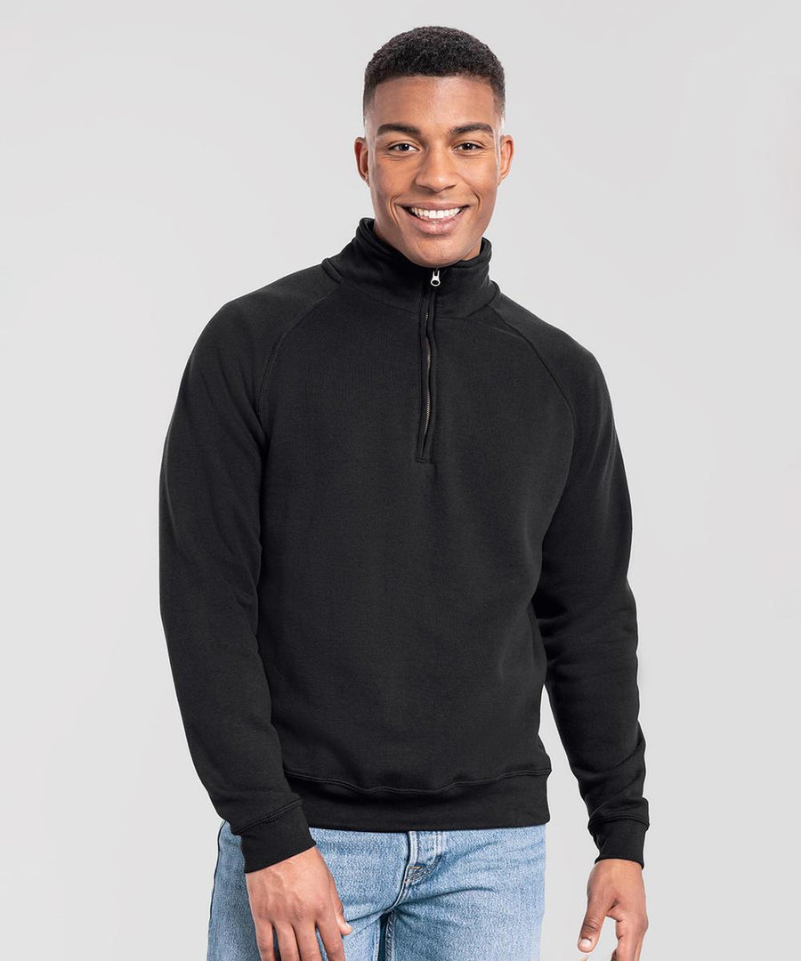 A person stands facing the camera, smiling, wearing a Fruit of the Loom Iconic premium zip-neck sweatshirt in black and light blue jeans against a plain light grey background.