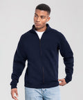 A person models the Fruit of the Loom Iconic premium sweat jacket in navy blue, worn over a white shirt and light blue jeans, standing against a plain light grey background and gazing to the side with one hand in their pocket.