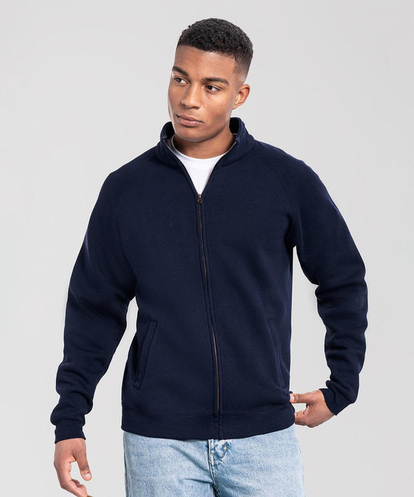 A person models the Fruit of the Loom Iconic premium sweat jacket in navy blue, worn over a white shirt and light blue jeans, standing against a plain light grey background and gazing to the side with one hand in their pocket.