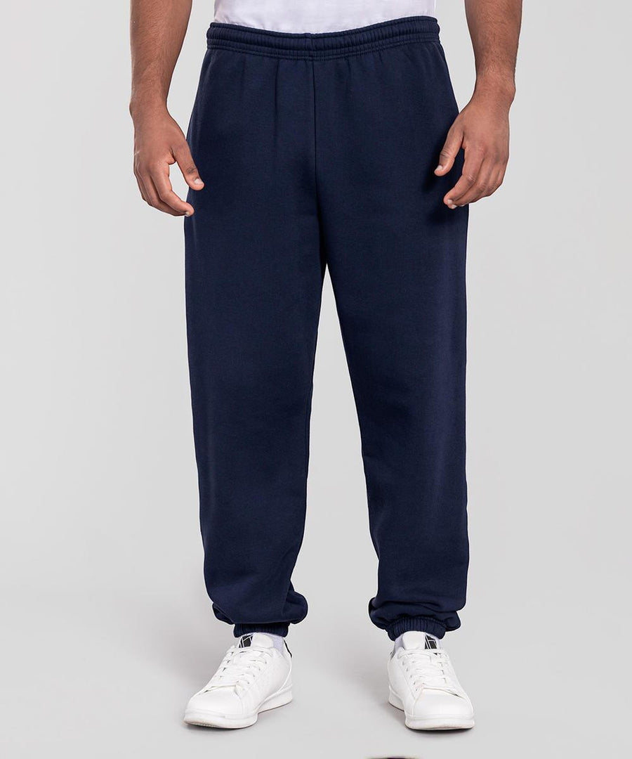 A person models Fruit of the Loom's Iconic premium elasticated cuff joggers in navy blue, paired with white trainers, standing against a plain light grey background. The photo is cropped from shoulders down, with hands relaxed at their sides.