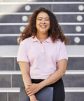 A smiling person with curly hair in a pink Fruit of the Loom SS505 shirt holds a black folder while standing in front of steps.