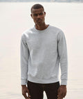 A man stands outdoors wearing a plain gray Fruit of the Loom SS800 sweatshirt and dark jeans, with a blurred background of water and landscape.