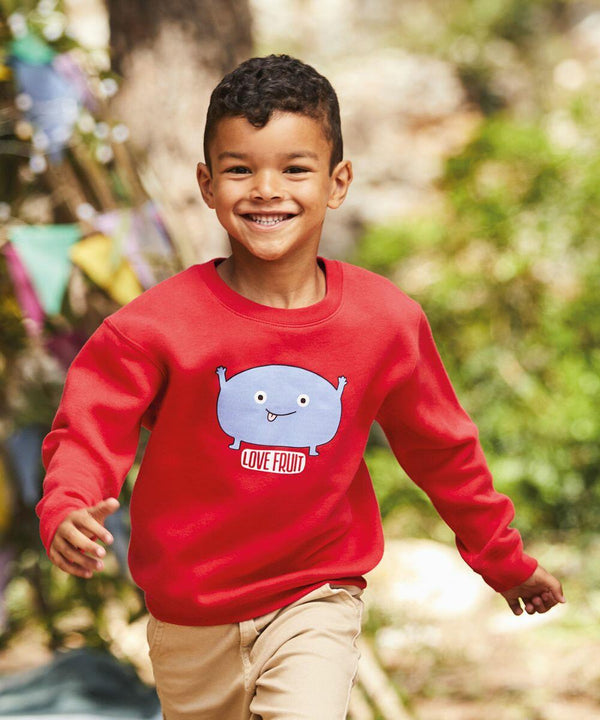 A young boy with short curly hair runs outside, smiling. He is wearing a red sweatshirt with a cartoon fruit character and the text "LOVE FRUIT" on it. The sweatshirt is a Fruit of the Loom SS801. Trees and colorful decorations are in the background.