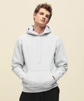 A person with short curly hair wearing a gray Fruit of the Loom SS824 hooded sweatshirt with their hands in the front pocket stands against a plain background.
