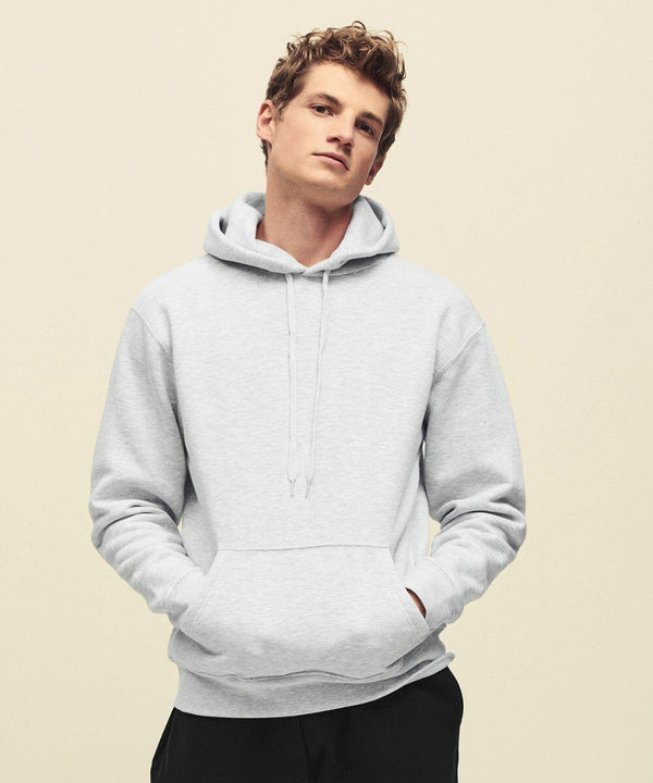 A person with short curly hair wearing a gray Fruit of the Loom SS824 hooded sweatshirt with their hands in the front pocket stands against a plain background.