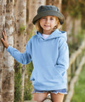 A young girl wearing a light blue Fruit of the Loom SS873 and a green hat stands by a tree, smiling and with her hands in her hoodie pocket. She is outdoors on a sunny day.
